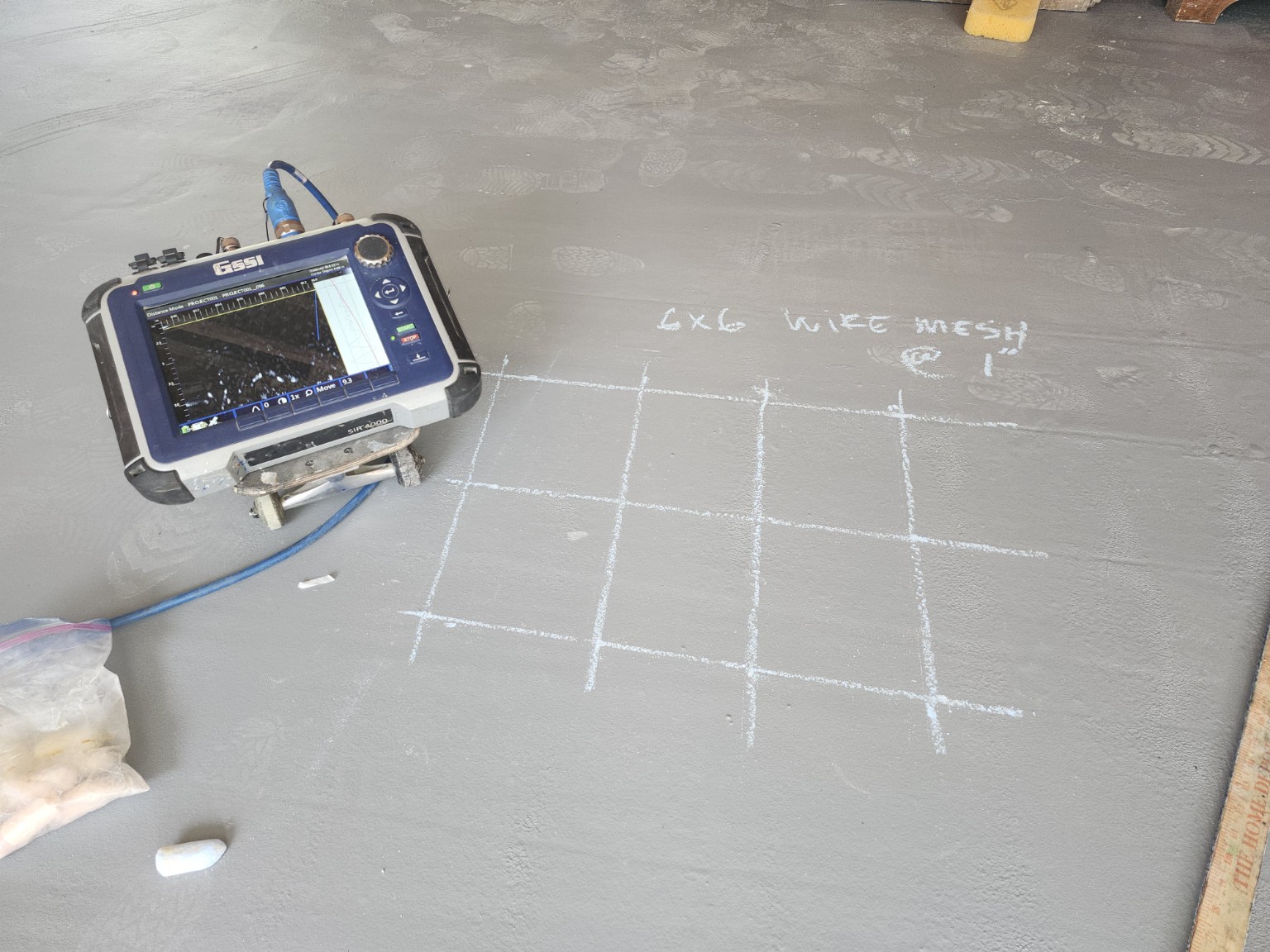 A GSSI SIR 4000 ground-penetrating radar unit on a freshly placed concrete topping beside a chalk grid labeled 6x6 wire mesh at 1 inch.